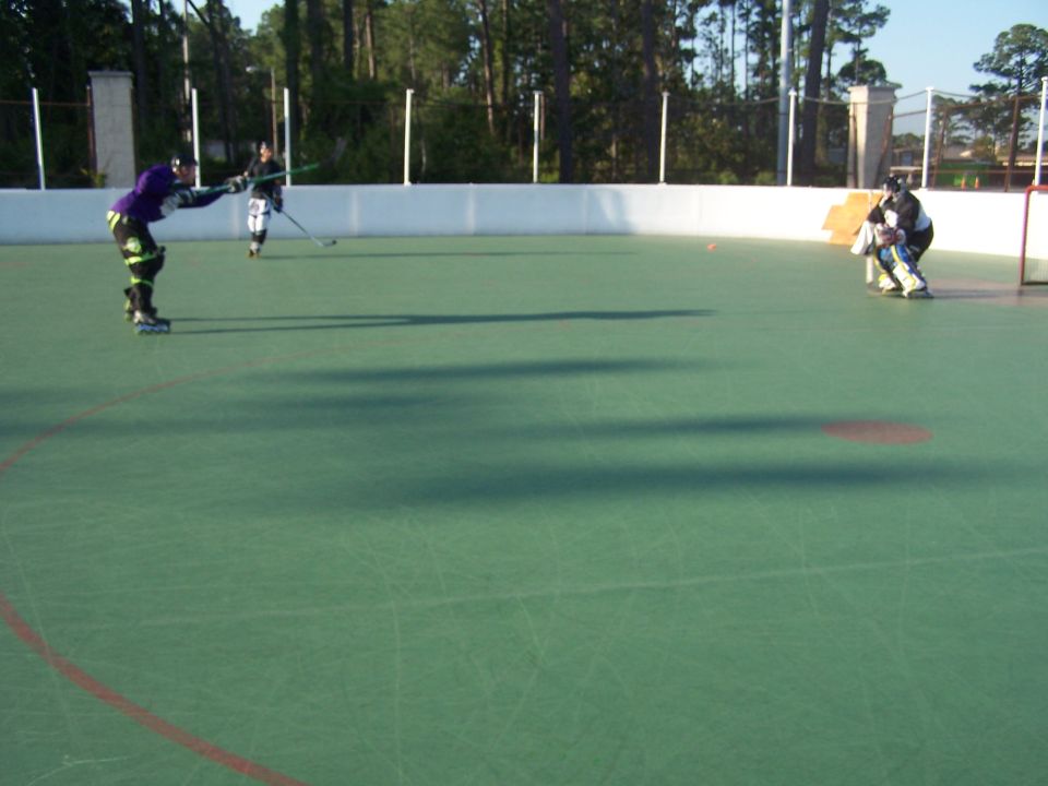 Roller hockey players on a green court with trees and fence in background. Supermotors.net logo at bottom right