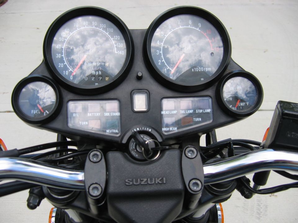Motorcycle dashboard with speedometer, tachometer, fuel gauge, oil temperature gauge, and indicator lights. Suzuki logo visible