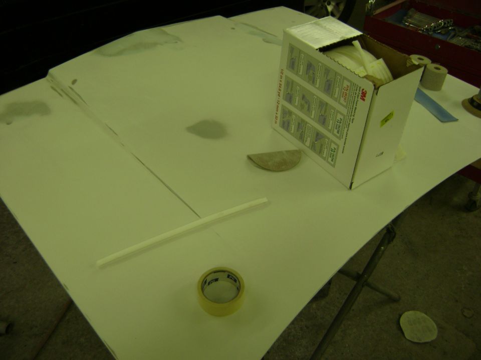 Car hood covered in white primer with a box of 3M masking tape, sandpaper, and a roll of tape