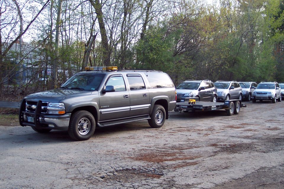 Gray SUV with yellow lights on top tows a trailer carrying two cars. More cars are parked behind it