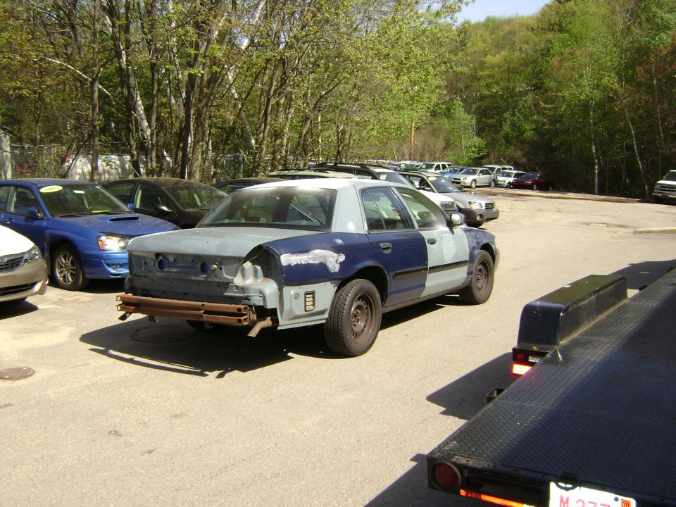 2001 Ford Crown Victoria with primer gray and blue mismatched body panels, missing rear bumper, parked in lot with other cars