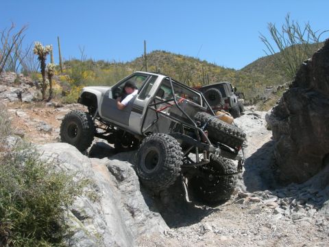 1986 Toyota Hilux - A heavily modified pickup truck with a roll cage and oversized tires tackles a challenging rocky terrain in a desert landscape.