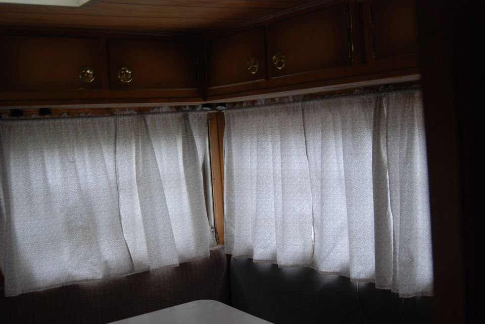 Camper interior with wood overhead cabinets and white curtains over corner windows