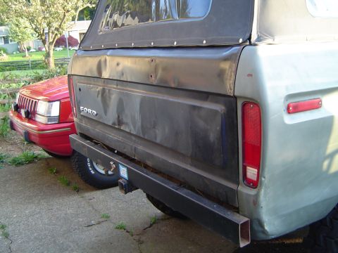 1978 Ford Bronco - Rear view of a 1978 Ford Bronco, showing its tailgate panel and aftermarket bumper, with a red vehicle parked alongside.
