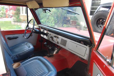 1977 Ford Bronco - The interior of a classic red Ford Bronco shows its dashboard, seats, and steering wheel, with a spare tire visible through the window.