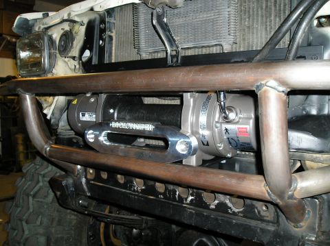 1986 Toyota Hilux - Close-up of a Warn electric winch mounted behind a bare tubular steel front bumper on a truck under construction in a garage, with the winc…