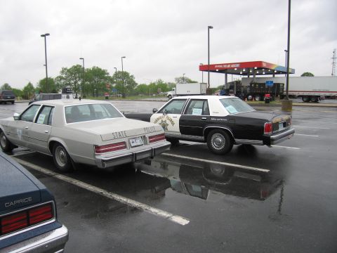 1991 Ford LTD Crown Victoria - silver State Police sedan and a black-and-white police cruiser parked on a rainy lot with a gas station and trucks in the background.