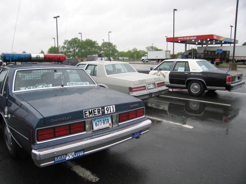 1991 Ford LTD Crown Victoria - Rear of a dark blue Chevrolet Caprice marked EMER 911 with light bar, parked among other police cars on a wet lot.