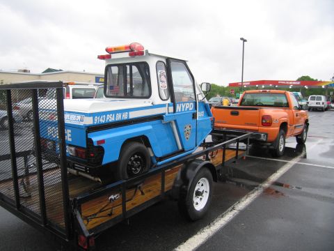 1991 Ford LTD Crown Victoria - blue and white NYPD three-wheeled scooter strapped to a utility trailer towed behind an orange pickup truck.
