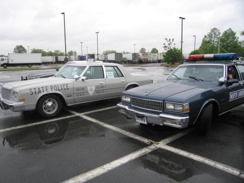 1991 Ford LTD Crown Victoria - silver Rhode Island State Police sedan parked next to a dark blue Chevrolet Caprice police cruiser on a wet lot.