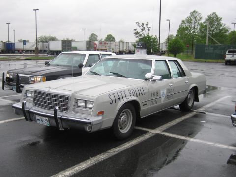 1991 Ford LTD Crown Victoria - silver Rhode Island State Police sedan with chrome push bar and roof light parked on a rainy lot beside a black car.