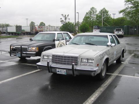 1991 Ford LTD Crown Victoria - silver State Police sedan and a black sedan with chrome push bar parked side by side on a rainy parking lot.