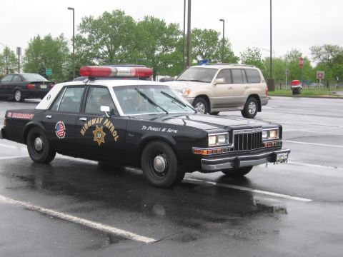1991 Ford LTD Crown Victoria - black-and-white Highway Patrol sedan with red roof light bar and star emblem parked on a wet lot, SUV behind.