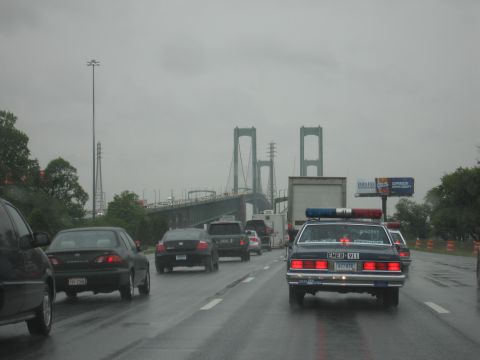 1991 Ford LTD Crown Victoria - dark police cruiser drives on a wet multi-lane highway toward a large green suspension bridge under gray skies.