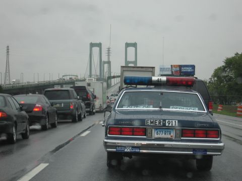 1991 Ford LTD Crown Victoria - Rear view of a blue Chevrolet Caprice EMER 911 cruiser with light bar driving on a wet highway toward a suspension bridge.
