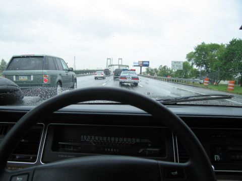 1991 Ford LTD Crown Victoria - Driver's view over a steering wheel and dashboard at a police car on a wet highway approaching a suspension bridge.