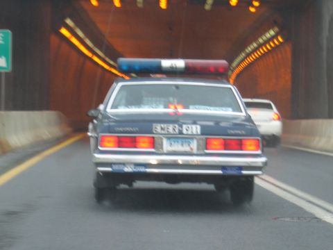 1991 Ford LTD Crown Victoria - Rear view of a dark Chevrolet Caprice EMER 911 cruiser with light bar driving inside an illuminated highway tunnel.