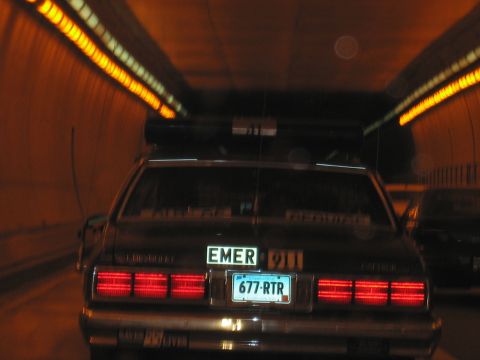1991 Ford LTD Crown Victoria - Rear of a dark police car with glowing taillights and EMER 677-RTR plate driving through a dimly lit tunnel.