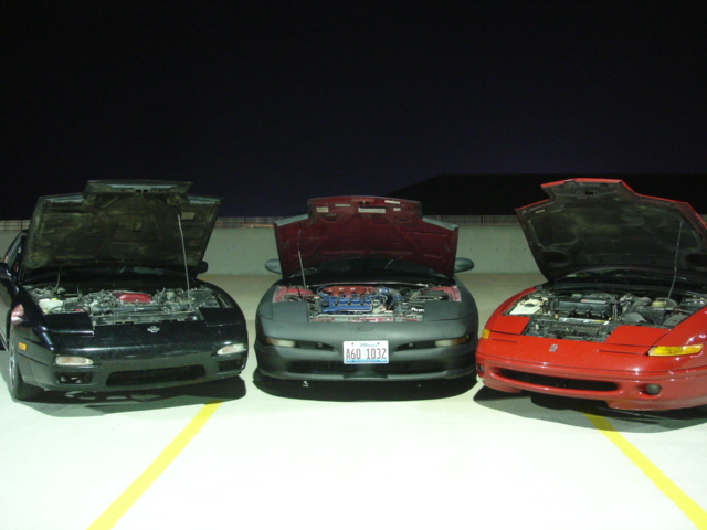 Three sports coupes, two dark and one red, with hoods open, parked in a lot at night
