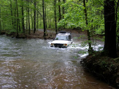 1993 Ford Bronco - A white Land Rover Discovery is driving through a shallow river in a forest. The vehicle has a roof rack and is kicking up water as it move…