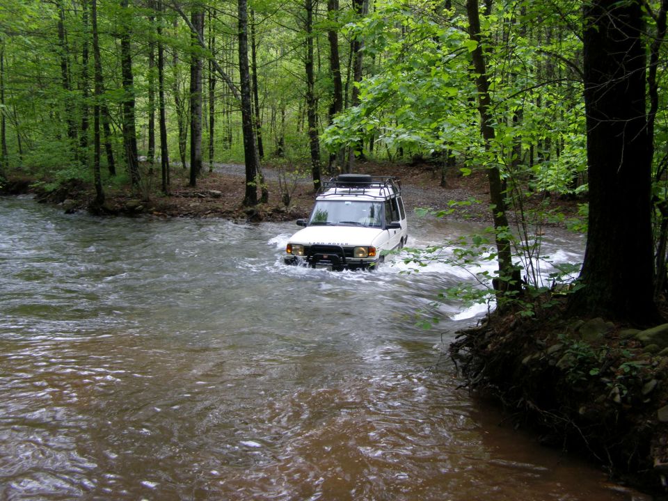White Land Rover Discovery driving through a river in a forest.