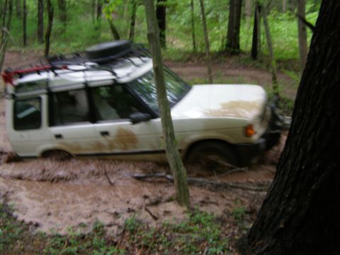 1993 Ford Bronco - A white SUV navigates a muddy trail through a dense forest, with its tires deep in the mud and mud splattered on its body.