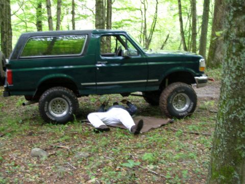 1993 Ford Bronco - A person in white pants works underneath the chassis of a dark green Ford Bronco parked in a wooded area. The vehicle is lifted on large ti…