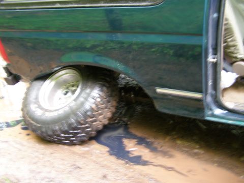 1993 Ford Bronco - The rear passenger side of a dark green SUV is partially submerged in mud, with its tire stuck in the muck. The vehicle's door is open, rev…