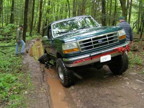 1993 Ford Bronco - A green Ford Bronco is stuck in a muddy puddle on a wooded trail. A Hi-Lift jack is mounted to the front bumper, and two people are visible…
