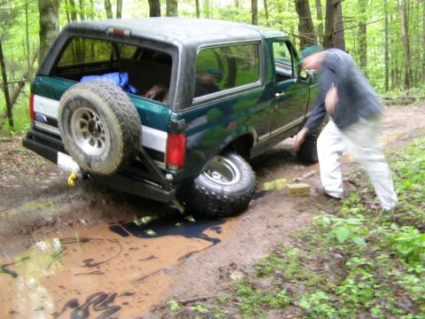 1993 Ford Bronco - A dark green Ford Bronco is stuck in a muddy puddle off-road, with its rear tire submerged. A person in light-colored pants is visible near…