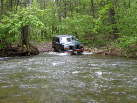 1993 Ford Bronco - A dark green Ford Bronco is driving through a shallow river in a wooded area. The vehicle is partially submerged in the water, with waves s…