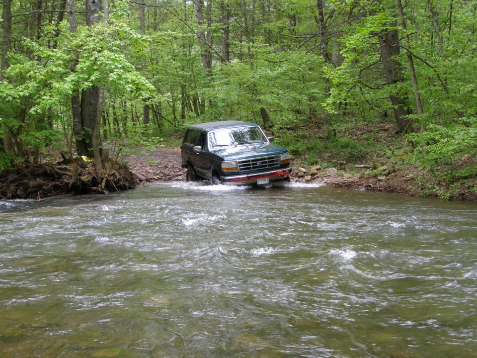 Dark green Ford Bronco driving through a river in a forest.