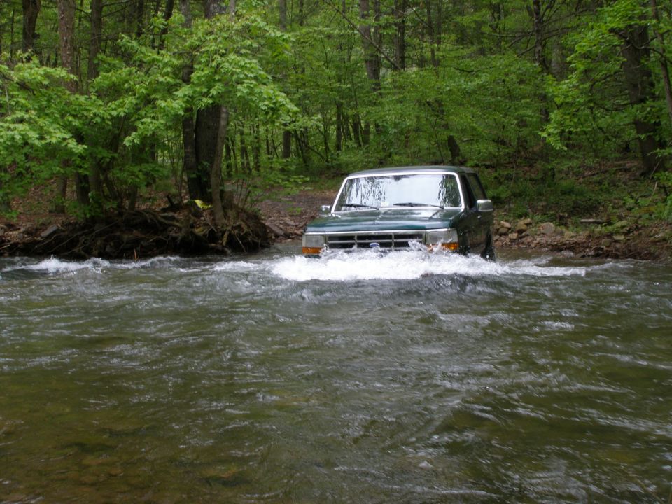 Green Ford Bronco driving through a forest river.