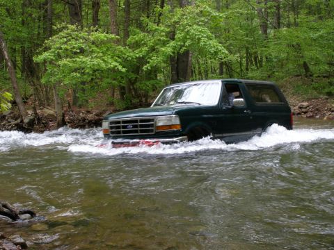 1993 Ford Bronco - A dark green Ford Bronco is driving through a shallow, rocky river surrounded by lush green trees.