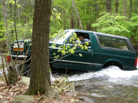1993 Ford Bronco - A dark green Ford Bronco is driving through a shallow stream in a wooded area. The vehicle is kicking up a lot of water as it moves forward.