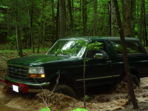 1993 Ford Bronco - A dark green 1993 Ford Bronco is driving through a muddy trail in a forest. The vehicle is partially submerged in brown water, with mud spl…
