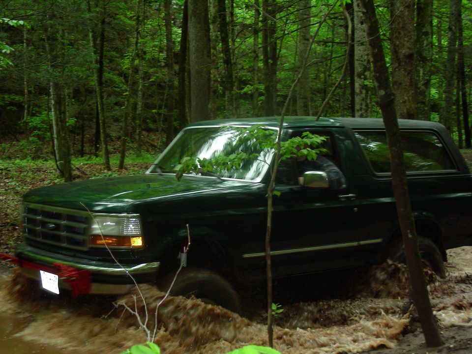 1993 Ford Bronco driving through mud and water in a forest.