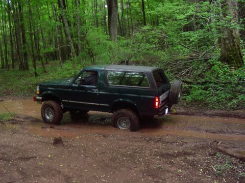 1993 Ford Bronco - A dark green Ford Bronco is driving through a muddy trail in a forest. The vehicle is angled to the right, showing its side profile as it n…