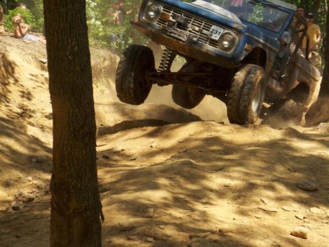 1990 Ford Bronco - A blue Ford Bronco is captured mid-air, its front tires lifted off the ground as it navigates a steep, dusty off-road trail. Spectators wat…