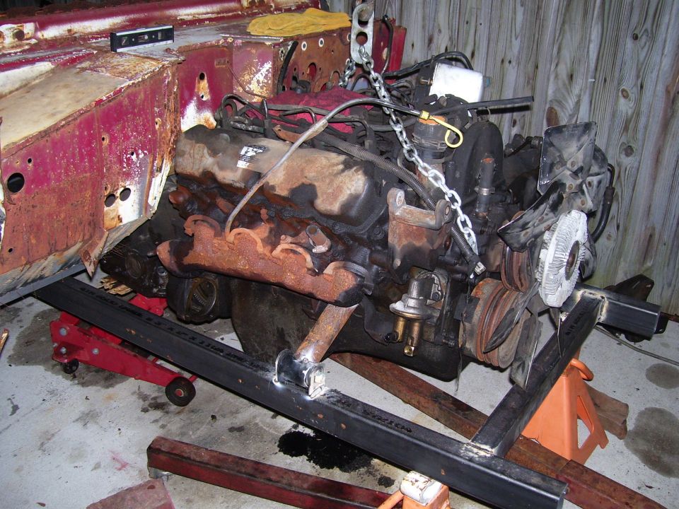 V8 engine with rusty exhaust manifolds on a black engine stand in a garage
