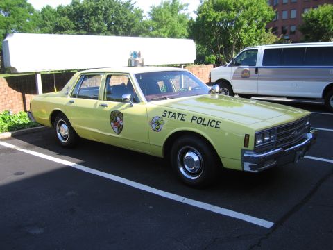 1991 Ford LTD Crown Victoria - pale yellow Maryland State Police Chevrolet Caprice sedan with shield emblem parked beside a van on a lot.