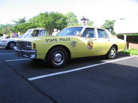 1991 Ford LTD Crown Victoria - pale yellow Maryland State Police Chevrolet Caprice sedan with shield emblem parked on asphalt with onlookers behind.