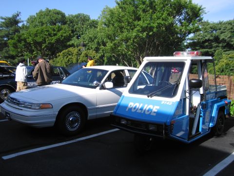 1991 Ford LTD Crown Victoria - blue NYPD three-wheeled police scooter parked beside a white Ford Crown Victoria police car under trees.
