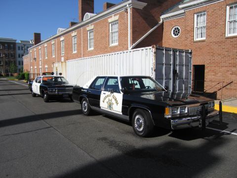 1991 Ford LTD Crown Victoria - black-and-white Highway Patrol Ford sedan and another police car parked beside a brick building and shipping container.