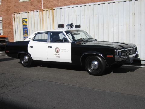 1991 Ford LTD Crown Victoria - black-and-white LAPD police sedan with twin roof lights and badge emblem parked beside a white fence in sunlight.