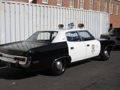 1991 Ford LTD Crown Victoria - Rear quarter of a black-and-white LAPD police sedan with twin roof spotlights parked by a white fence and brick building.