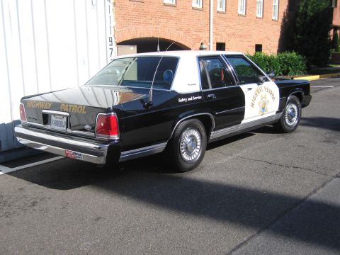 1991 Ford LTD Crown Victoria - black-and-white Highway Patrol police sedan parked beside a shipping container, shown from the rear three-quarter angle.
