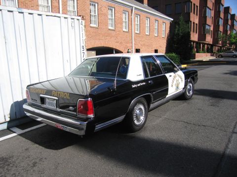 1991 Ford LTD Crown Victoria - black-and-white Highway Patrol sedan with chrome wire wheels parked in front of a brick building, viewed from the rear.