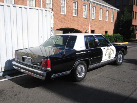 1991 Ford LTD Crown Victoria - black-and-white Highway Patrol police sedan parked near a shipping container and brick building, seen from the rear quarter.