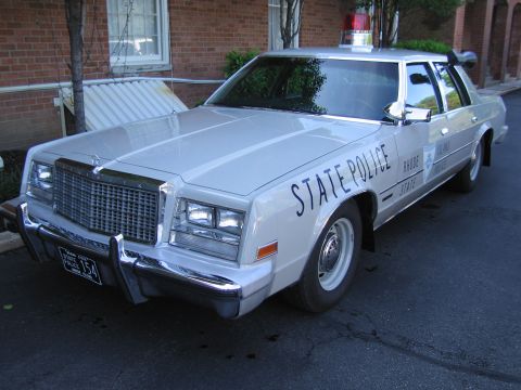 1991 Ford LTD Crown Victoria - white Chrysler State Police sedan with a roof lightbar and push bar, parked beside a brick building.